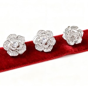 Puede incluir: Tres pendientes plateados con forma de flor y detalles de cristal transparente. Los pendientes están colocados sobre una superficie de terciopelo rojo. Cada pendiente presenta un cristal central rodeado de cristales más pequeños.