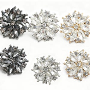 May include: Six flower-shaped rhinestone buttons. Two are dark gray, two are clear, and two are gold. Each button has a central cluster of small rhinestones surrounded by larger, petal-shaped rhinestones.