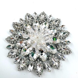 Puede incluir: Broche grande y brillante de cristal transparente y plateado. La broche circular presenta un grupo central de cristales transparentes, rodeado de piedras claras más grandes y facetadas. El diseño es intrincado y llamativo.