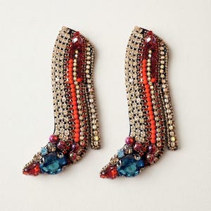 Op de afbeelding: Een paar broches in de vorm van schoenen met hoge hakken, versierd met rijen heldere kristallen, oranje kralen en donkerrode accenten. De neus is voorzien van een grote blauwe gefacetteerde steen, kleine rode kristallen en metalen kralen.