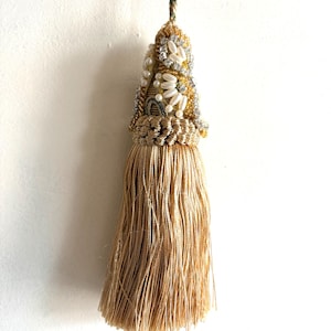 Puede incluir: Una borla decorativa con largos flecos de color oro claro suspendida de un cordón trenzado. La sección superior está adornada con perlas blancas de imitación, cristales transparentes y cuentas doradas en un patrón denso.