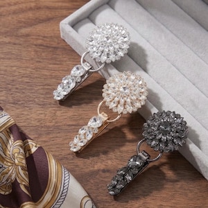 Può includere: Tre clip in metallo con teste circolari a forma di fiore incastonate con cristalli trasparenti, color champagne e grigio scuro. Ogni clip presenta un meccanismo a cerniera e uno stelo decorato con pietre di cristallo sfaccettate.
