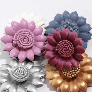 May include: A collection of five layered flower-shaped ornaments crafted from leather. The flowers feature pointed petals in white, light pink, dark pink, blue, silver, and gold, each with a textured, coiled center made of small leather strips.