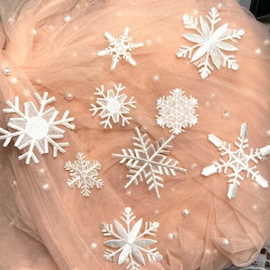 以下が含まれることがあります： ベージュのチュール生地に白い雪の結晶の装飾が施されたクローズアップ。雪の結晶は様々なサイズとスタイルで、中には他のものよりも複雑なデザインのものもある