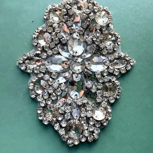 May include: A decorative applique featuring a dense arrangement of clear, faceted rhinestones and crystals in various shapes, including round, oval, and marquise, set against a light-colored fabric backing in an elongated, symmetrical floral-inspired pattern.