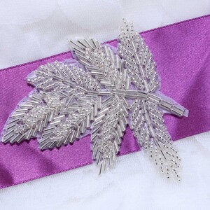 May include: A silver beaded leaf applique with a purple satin ribbon. The leaf is made of small, silver beads that are sewn together to create a delicate and intricate design.