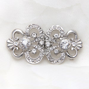 May include: A silver-toned metal cloak clasp featuring two symmetrical, ornate scrollwork pieces. Each side is embellished with numerous small, clear, round-cut rhinestones and a larger, prominent, central clear rhinestone set in the middle.