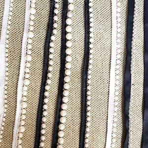 May include: Close-up of various decorative ribbons and trims. The image features white, black, and cream-colored ribbons with pearl accents and satin finishes. The ribbons are arranged vertically against a textured background.