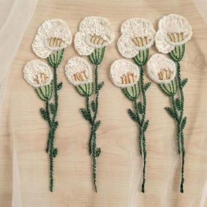 Könnte beinhalten: Vier florale Applikationen mit weißen Paillettenblütenblättern, goldfarbenen Perlenzentren sowie grünen, perlenbesetzten Stielen und Blättern. Jede Applikation zeigt eine langstielige Blume mit runder Blüte aus detaillierter Perlen- und Paillettenstickerei.