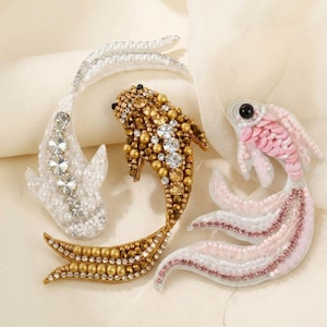 May include: Three decorative fish-shaped brooches crafted with intricate beadwork. One is white with clear crystals, one is gold with metallic beads and crystals, and one is light pink with sequins and a black eye bead, all featuring curved tails.