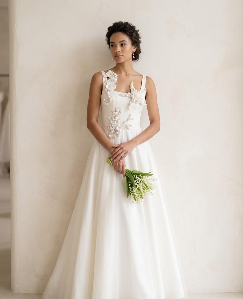 A model in a white bridal gown embellished with pearl seahorse, starfish, and coral appliques. A luxury white wedding dress with coastal themed pearl appliques on the bodice and shoulder.