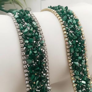Può includere: Due braccialetti con una fascia centrale di pietre verde scuro di forma irregolare. Un braccialetto ha un bordo color argento con piccole pietre trasparenti, l'altro ha un bordo color oro con pietre trasparenti simili.