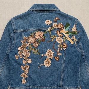 May include: A blue denim jacket featuring intricate embroidery on the back. The design displays a bird with teal and tan wings perched on brown branches adorned with light tan and brown flowers and green leaves, extending across the back panel.