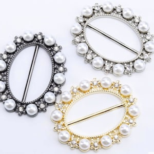 以下が含まれることがあります： 装飾的なデザインの楕円形の金属製のバックルが3つ。各バックルは、フェイクパールとラインストーンの列で飾られています。バックルはシルバー、ゴールド、ブラックです。