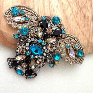 Peut inclure: Broche ornée de pierres précieuses scintillantes, composée de teintes turquoise, noires, transparentes et champagne. La broche, bordée d'un filigrane doré, est posée sur une surface en bois clair.