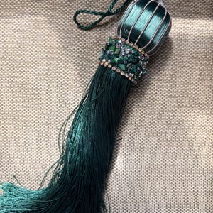 May include: An emerald green tassel with a beaded accent and a looped cord. The tassel has a ribbed, cylindrical top and a long, flowing fringe. The accent includes green stones and clear rhinestones.