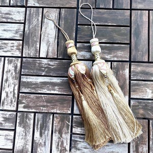 May include: Two decorative tassels with gold and white fringe. The tassels have a pink and white patterned design and are tied with a loop.