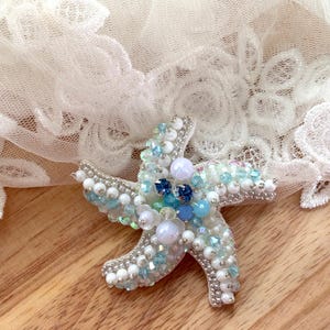 Puede incluir: Un broche de estrella de mar hecho a mano, adornado con cuentas blancas, azul claro y azul oscuro. El broche tiene una base de metal plateado y está decorado con cuentas de varios tamaños. El broche es un accesorio decorativo.