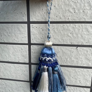 Puede incluir: Un borlón azul y blanco con una parte superior de cuentas decorativas. El borlón tiene tres hebras de flecos, dos azules y una blanca.