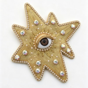 Könnte beinhalten: Goldene sternförmige Brosche mit zentralem Augendesign. Die Brosche hat ein braunes Auge, umgeben von goldenen Strahlen, Perlen und schillernden Kristallen. Die Ränder des Sterns sind mit kleinen, klaren Kristallen und Goldfaden besetzt.