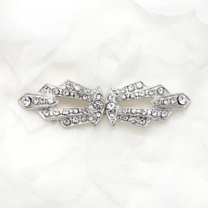 May include: A silver-toned brooch with a geometric design, featuring a central motif of two interlocking ovals. The brooch is adorned with numerous clear rhinestones.