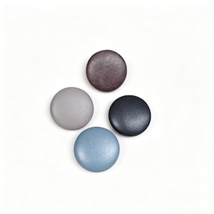 May include: Four circular buttons with a textured surface arranged in a cluster. The buttons are coloured in shades of dark brown, light grey, dark navy blue, and light blue. Each button features a smooth, rounded top and a consistent circular shape.