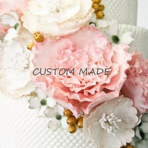 Puede incluir: Primer plano de un arreglo floral con grandes flores rosas y crema rizadas, pequeñas flores blancas y detalles dorados. El texto "CUSTOM MADE" es visible.