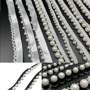May include: An assortment of decorative trims featuring white faux pearls. The trims include white satin ribbon, white lace, and black and white woven trim, all adorned with various sizes of faux pearls. The trims are arranged on a black surface.