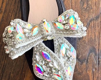 May include: A black pointed-toe flat shoe featuring a large decorative bow on the front. The bow is heavily embellished with various clear and iridescent rhinestones, crystals, and silver-toned beads, creating a highly reflective surface.