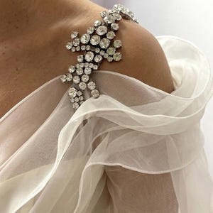 May include: A sparkling shoulder accessory with a floral design, adorned with numerous clear crystals. The crystals are set on a silver-toned metal base. The accessory is placed on a white, sheer fabric, creating a layered effect. This elegant piece is perfect for special occasions.