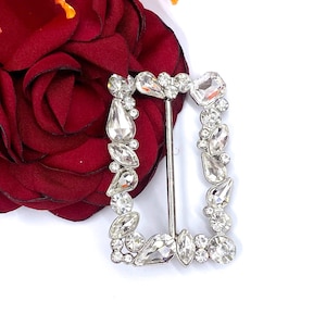 May include: A silver-toned buckle with a rectangular frame adorned with clear rhinestones. The buckle is designed with a variety of rhinestone shapes, including round, oval, and teardrop.