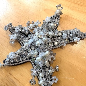 Puede incluir: Una aplicación en forma de estrella con una base negra densamente cubierta de pedrería transparente facetada, perlas artificiales blancas y detalles de alambre plateado. El diseño incorpora racimos de cristales y perlas en forma de flor.