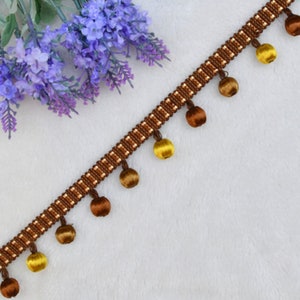 K&ouml;nnte beinhalten: Braune und goldene Franse mit einem sich wiederholenden Muster aus kleinen Quadraten. Die Franse hat eine Reihe von runden Fransen in Braun und Gold.
