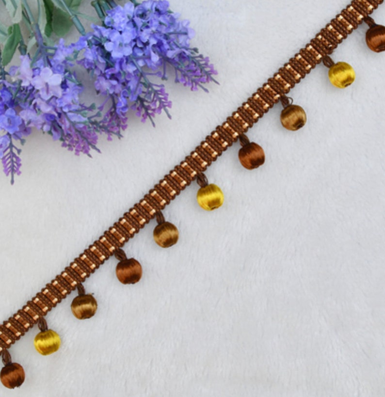 K&ouml;nnte beinhalten: Braune und goldene Franse mit einem sich wiederholenden Muster aus kleinen Quadraten. Die Franse hat eine Reihe von runden Fransen in Braun und Gold.