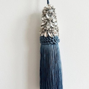 Peut inclure: Un pompon décoratif bleu doté d'une partie supérieure ornée de cristaux transparents à facettes et de petites perles blanches. Le pompon est suspendu à un cordon torsadé bleu assorti et se compose de nombreux fils bleus fins et droits.