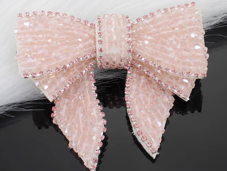 K&ouml;nnte beinhalten: Ein dekoratives Schleifen-Accessoire, das mit hellrosa facettierten Perlen bedeckt und an den R&auml;ndern mit Reihen kleiner rosa Strasssteine verziert ist. Die Mitte der Schleife hat ein vertikales Band aus passenden rosa Perlen und Strasssteinen.
