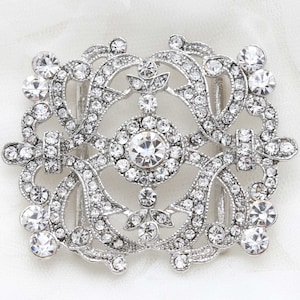 May include: Silver-toned, ornate buckle with a rectangular shape. The buckle is heavily embellished with clear, sparkling rhinestones of various sizes, arranged in a symmetrical, floral-inspired pattern. The design includes a central circular element and curved, openwork details.