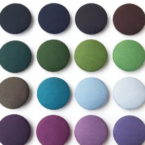 May include: Sixteen circular fabric-covered buttons in a variety of colours. The buttons include shades of navy, forest green, teal, light blue, and purple. The buttons are arranged in a grid pattern.
