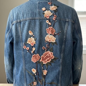 May include: A medium-wash blue denim jacket featuring an intricate embroidered floral design on the back. The embroidery depicts a branching stem with multiple blooming flowers in shades of soft pink, peach, and cream, accented by dark green leaves.