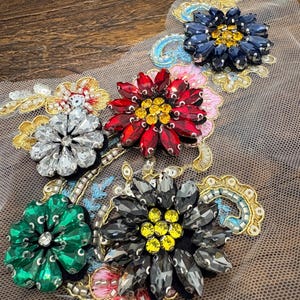 Puede incluir: Un aplique decorativo con múltiples formas florales hechas de cristales facetados en rojo, verde, transparente, azul oscuro y gris. Las flores están acentuadas con centros amarillos, bordados en hilo dorado y pequeñas lentejuelas sobre malla beige.