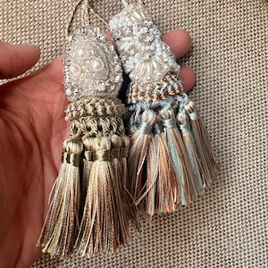 Può includere: Due nappe decorative con intricate lavorazioni di perline e ricami sulle sezioni superiori. La nappa sinistra ha fili di seta dorati e bronzo, mentre quella destra presenta toni azzurri, argento e rame con accenti di perle.