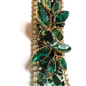 May include: A close-up of a decorative, rectangular object with a white base, adorned with emerald green gemstones, gold accents, and clear rhinestones. The design includes leaf-shaped embellishments and smaller green stones.