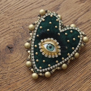 May include: A heart-shaped brooch with a dark green velvet centre, featuring a gold-rimmed eye and surrounded by rhinestones and gold beads. The brooch has a textured, fur-like appearance and is set against a wooden surface.