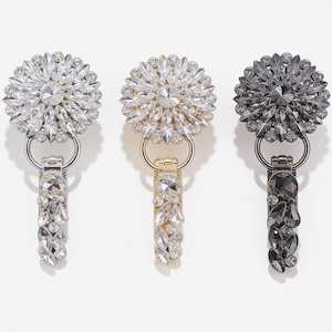 Può includere: Tre chiusure decorative con strass. Ogni chiusura presenta un design circolare con una pietra centrale circondata da pietre più piccole. Uno è argentato, uno è dorato e uno è grigio scuro, tutti con una sezione rettangolare pendente.