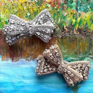 May include: Two decorative jeweled bows, one silver and one gold, with clear rhinestones, on a blue and green painted background.