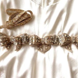 Puede incluir: Un cinturón de tono dorado con tres lazos decorativos adornados con cristales transparentes y cuentas metálicas. El cinturón incluye una cinta larga a juego de tono dorado con bordados intrincados y adornos de cristal a lo largo de toda su longitud.