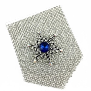 Peut inclure: Broche flocon de neige argentée avec une grosse pierre bleue ronde au centre et de petits cristaux transparents. La broche est posée sur une base rectangulaire recouverte de strass argentés, créant un effet scintillant. Idéale pour les tenues de soirée.