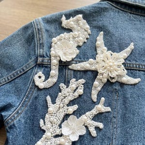 Pode incluir: Casaco de ganga azul adornado com aplicações brancas de cavalo-marinho, estrela-do-mar e coral. As aplicações apresentam contas semelhantes a pérolas e delicados detalhes de flores brancas, criando um tema náutico. O casaco tem uma lavagem azul clássica.