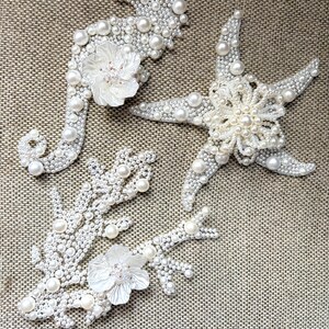 Flat lay image of three ocean themed pearl appliques showcasing the smooth plastic backing.

Handmade coastal pearl patches displayed on a glass plate with sand and real seashells.

A close up look at the shimmering iridescent beads and pearls on a handmade coral patch.

Creative DIY craft ideas displaying white pearl ocean motifs used for fashion and home decor.