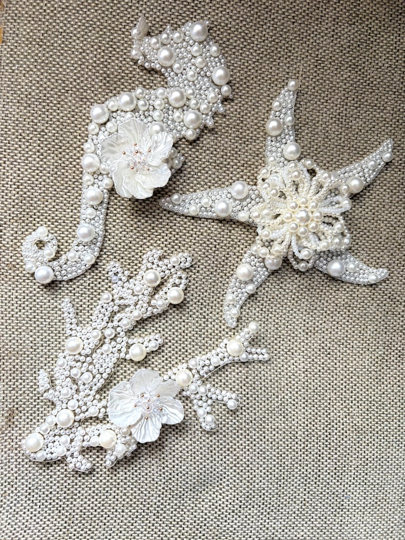 Flat lay image of three ocean themed pearl appliques showcasing the smooth plastic backing.

Handmade coastal pearl patches displayed on a glass plate with sand and real seashells.

A close up look at the shimmering iridescent beads and pearls on a handmade coral patch.

Creative DIY craft ideas displaying white pearl ocean motifs used for fashion and home decor.